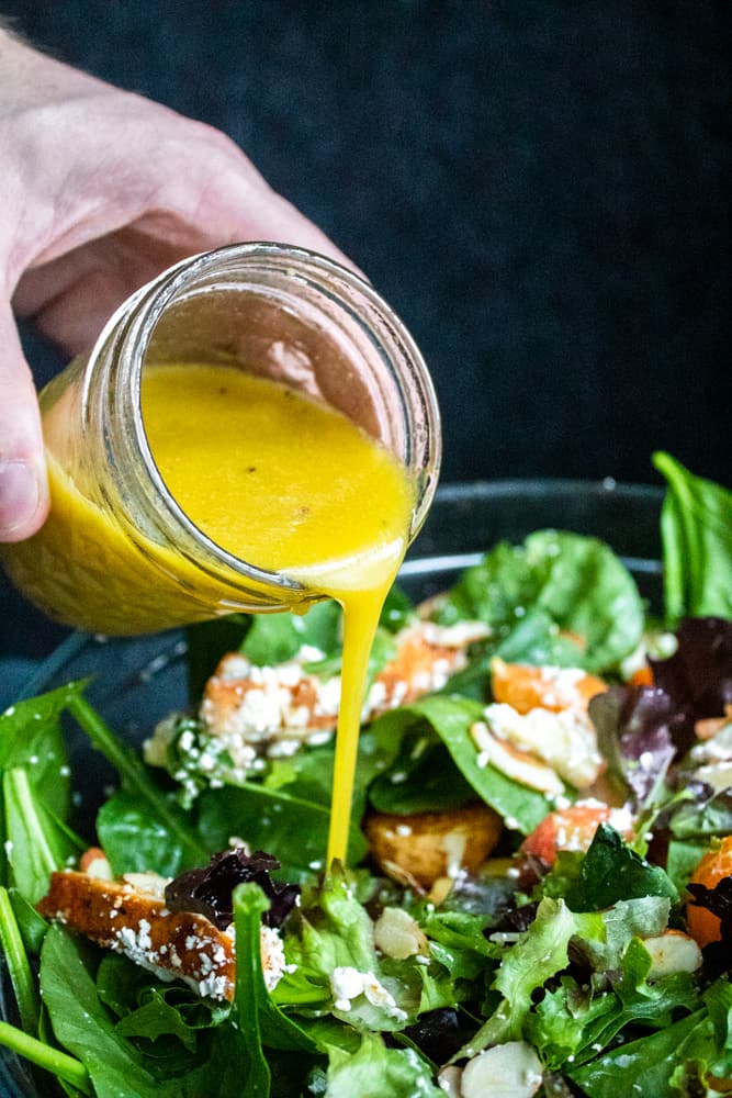 Hand pouring honey Dijon vinaigrette dressing over a glass bowl filled with salad.