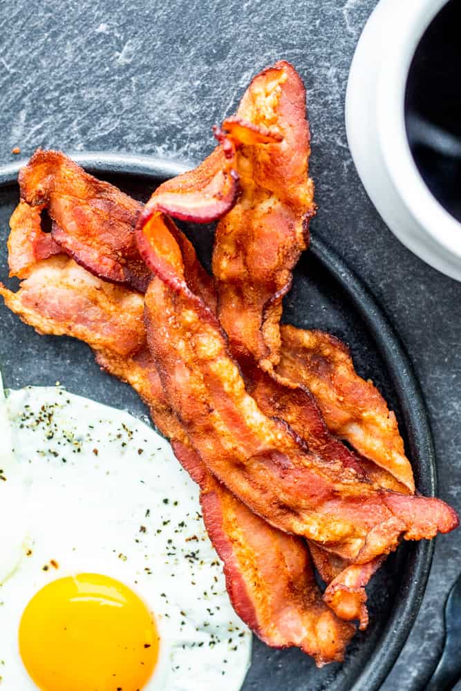 Air fryer bacon on a black plate with egg on the side.