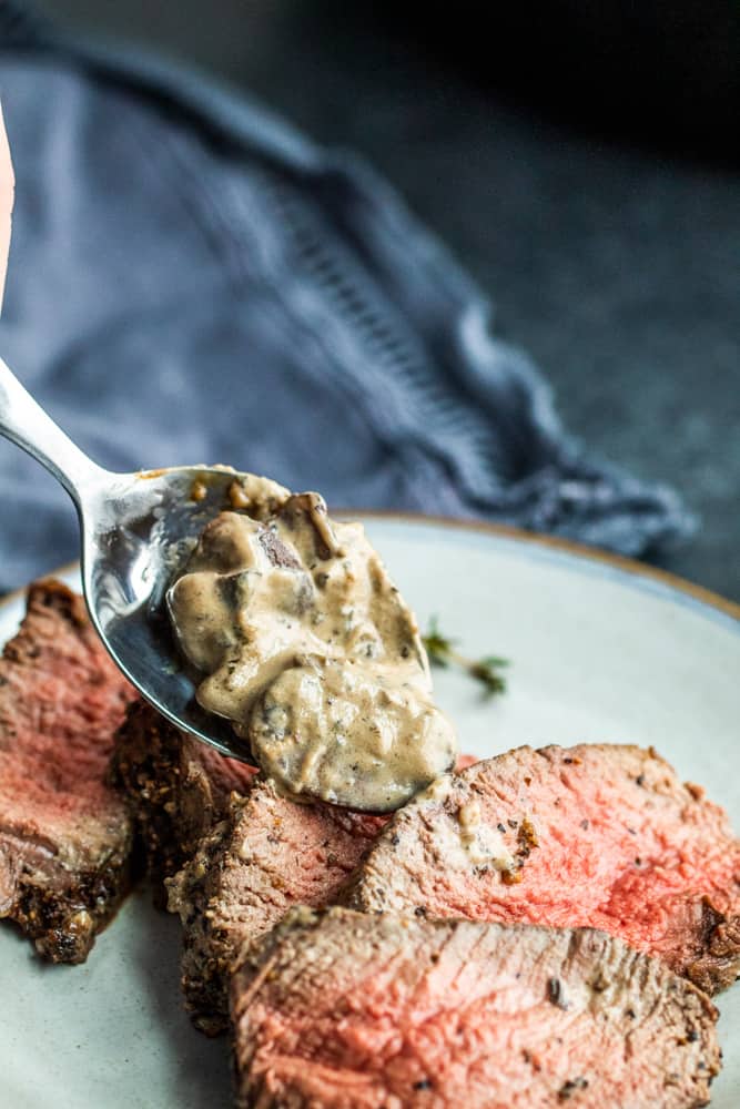 Silver spoon pouring mushroom sauce over sliced peppercorn steak on a gray plate.