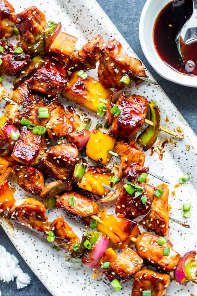 Overhead shot of teriyaki chicken skewers on a white platter with small bowl of teriyaki sauce on the side.