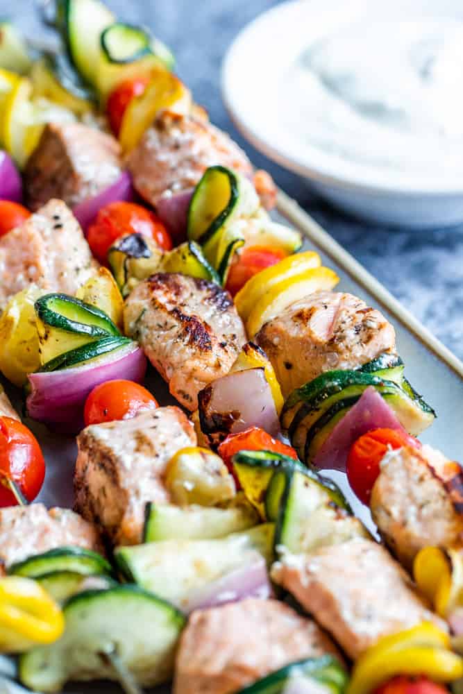 Angle shot of grilled salmon kabobs on a gray plate with sauce in the background.