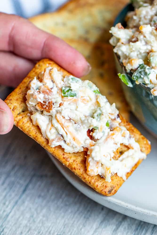 Hand picking up cracker with dip on top off white plate.