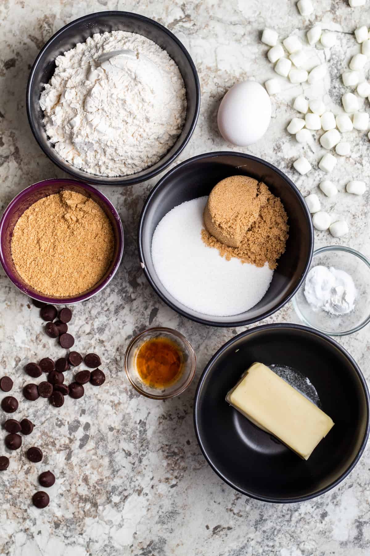 Overhead shot of s'mores cookie ingredients in small bowls on a white counter.