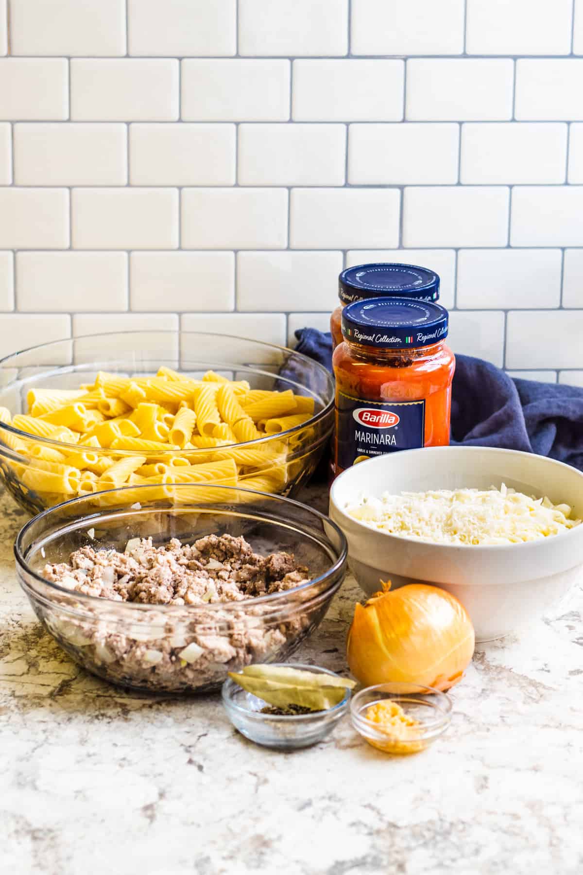 Mostaccioli ingredients in glass bowls laid out on a white counter.