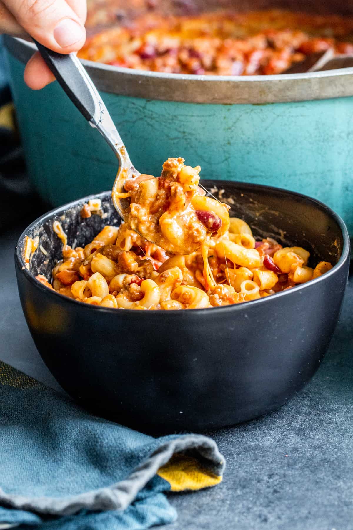 Spoon scooping up chili with noodles from black bowl.