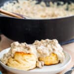 Two biscuits topped with gravy on a gray plate with skillet of gravy in background.