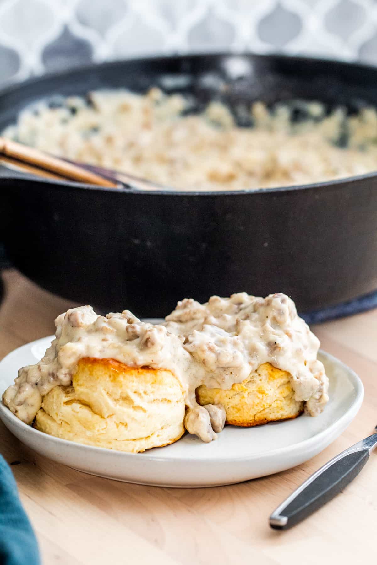 Two biscuits topped with gravy on a gray plate with skillet of gravy in background.