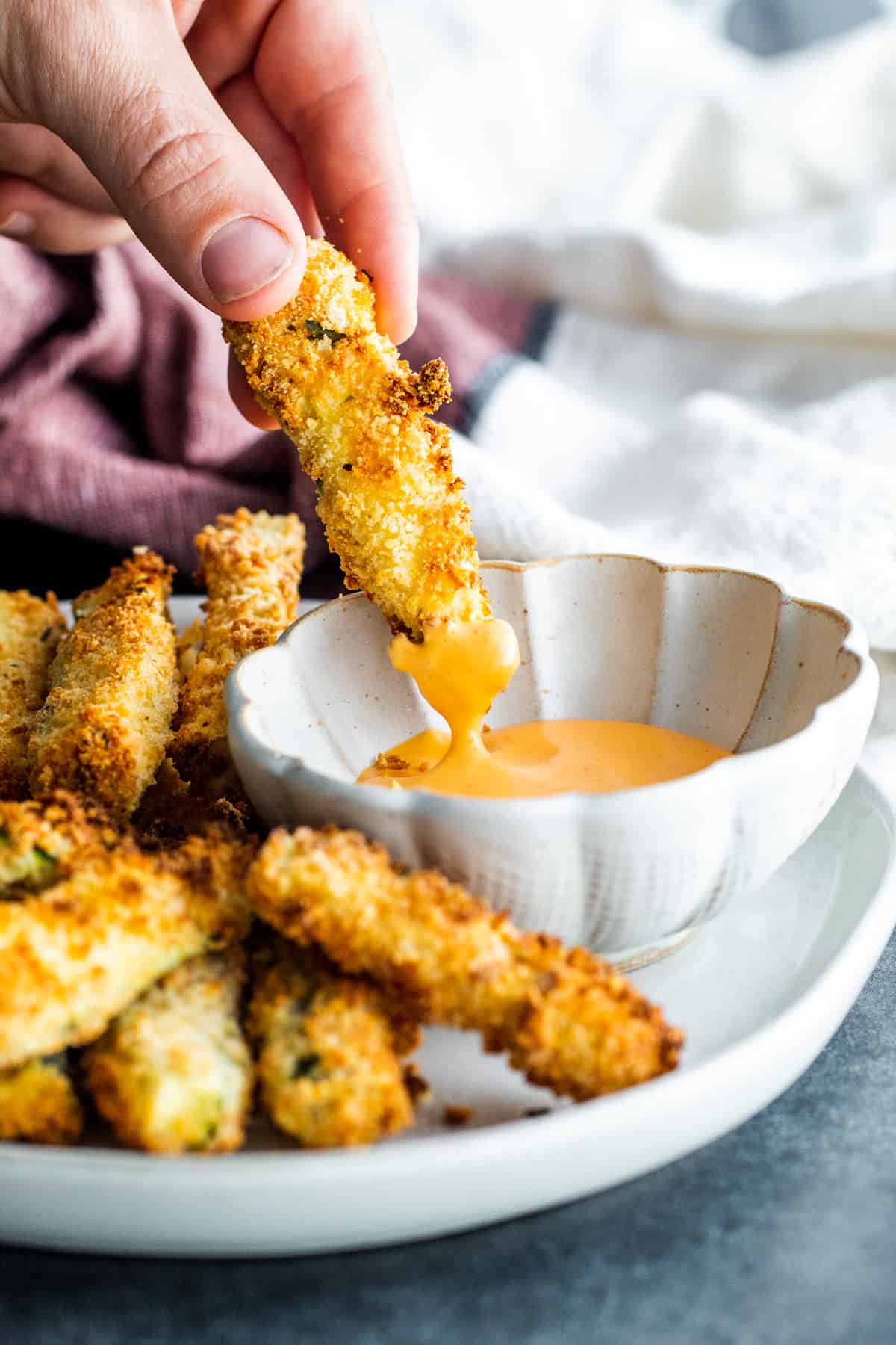 Hand dipping fried zucchini into a small white bowl of sauce.